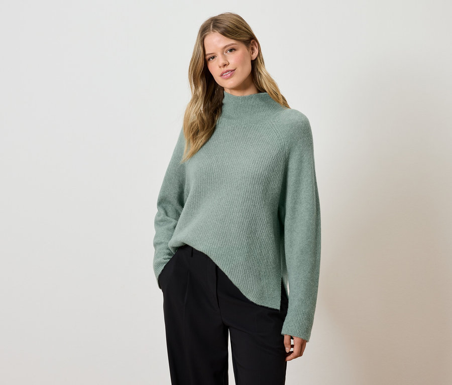 Frau posiert in grünem Pullover und schwarzer Hose.