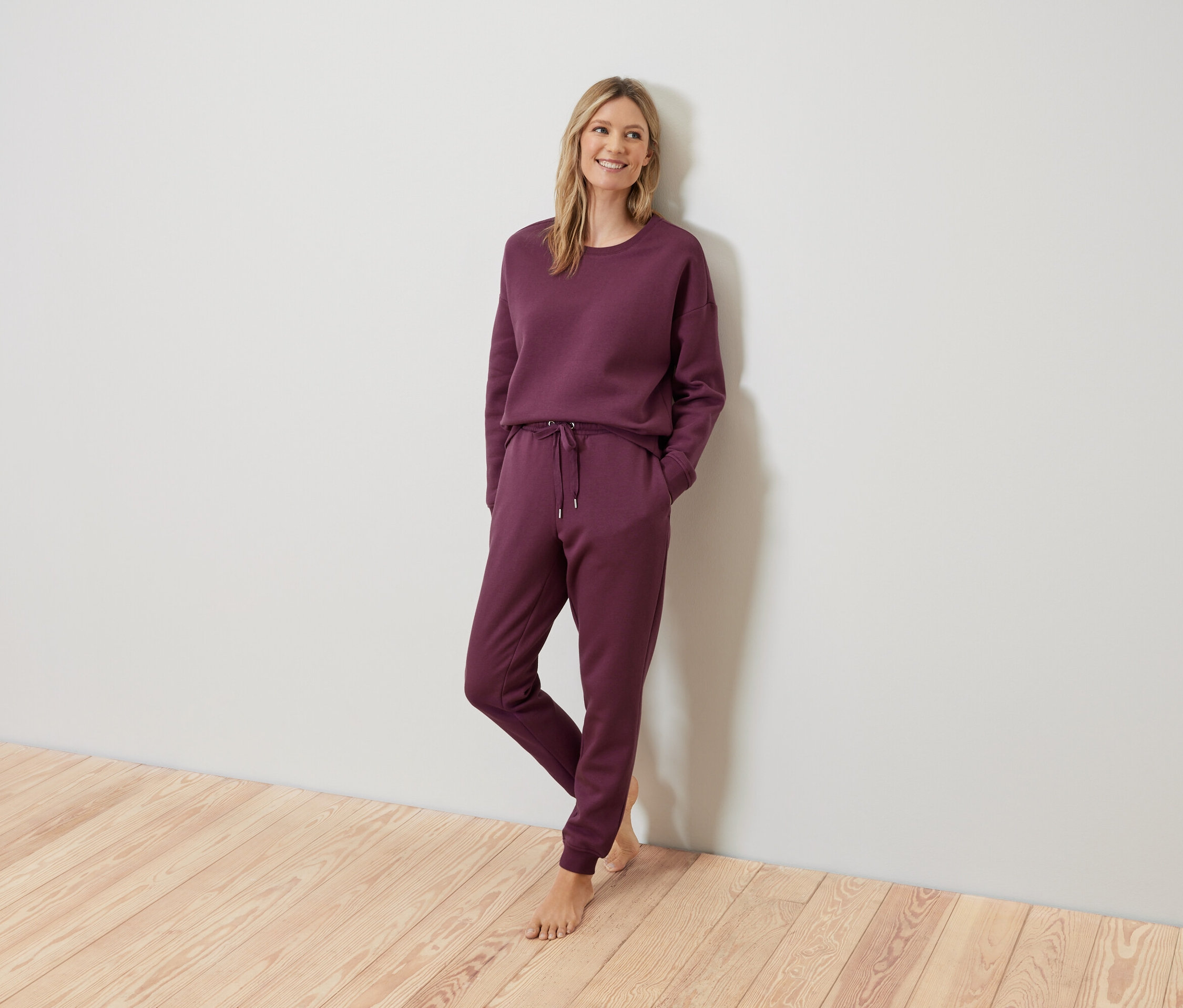 Blonde Frau posiert in einem burgunderroten Trainingsanzug an einer Wand.