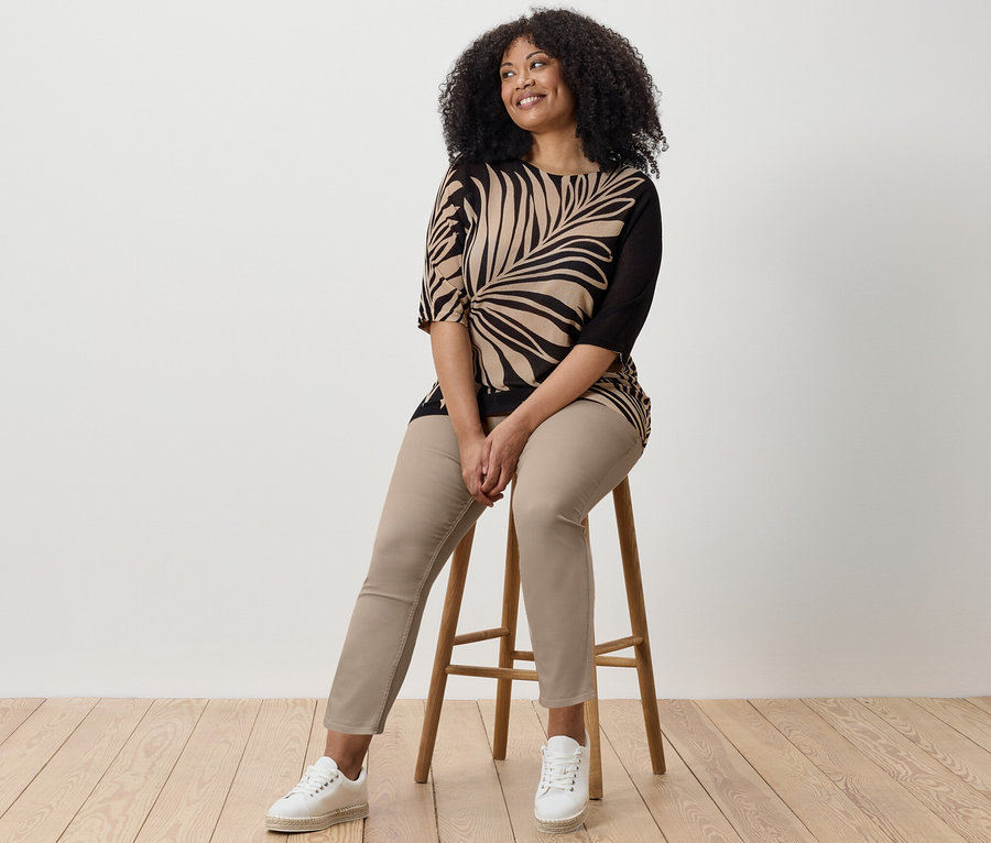 Frau sitzt auf einem Hocker und trägt 7/8-Treggings, ein bedrucktes Oberteil und Turnschuhe.