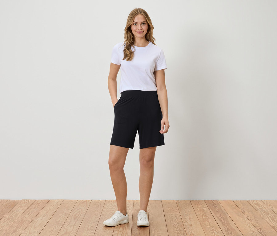 Frau mit weißem T-Shirt und schwarzen Shorts steht vor einer Wand.