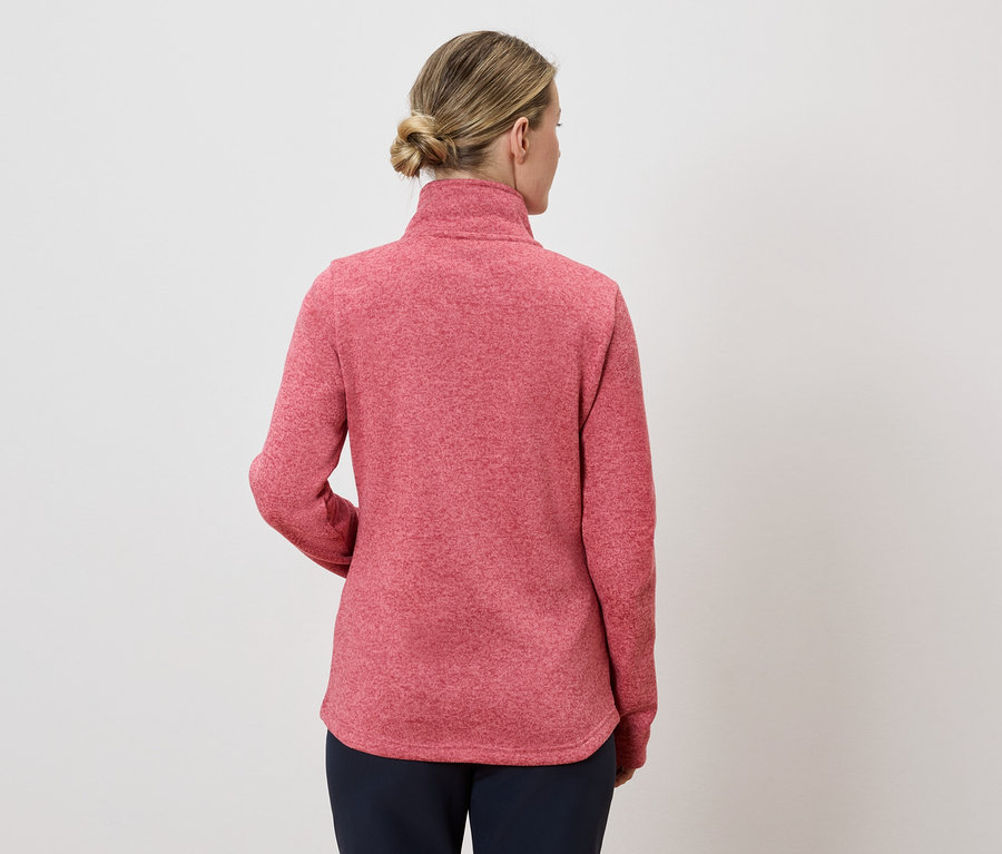 Rückansicht einer Frau mit blondem Haar, die einen roten Pullover und eine dunkle Hose vor einem weißen Hintergrund trägt.