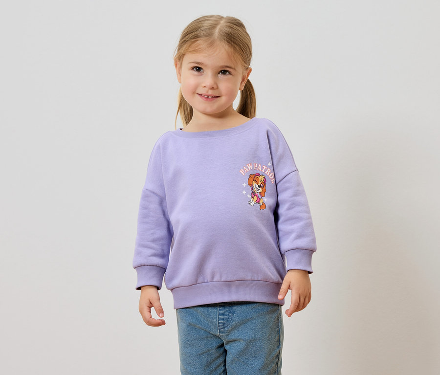 Mädchen mit Zöpfen trägt einen lila Pullover mit Paw Patrol Aufdruck und blaue Jeans.