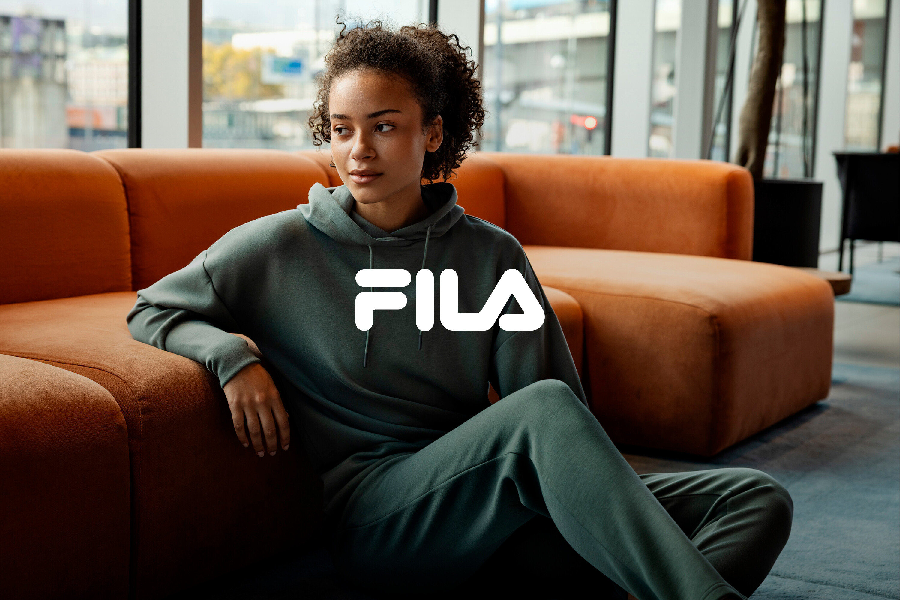 Eine Frau sitzt auf dem Boden, angelehnt an ein orangefarbenes Sofa, und trägt einen grünen Fila-Trainingsanzug.