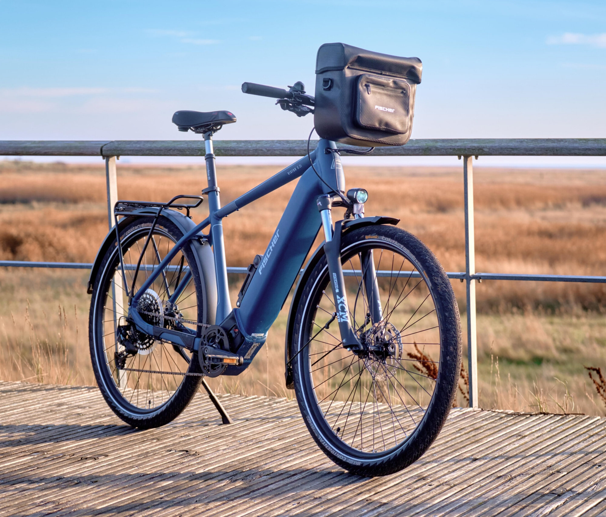 Blaues E-Bike mit FISCHER Lenkertasche PLUS+ Terra steht auf einem Holzsteg vor einer Dünenlandschaft.