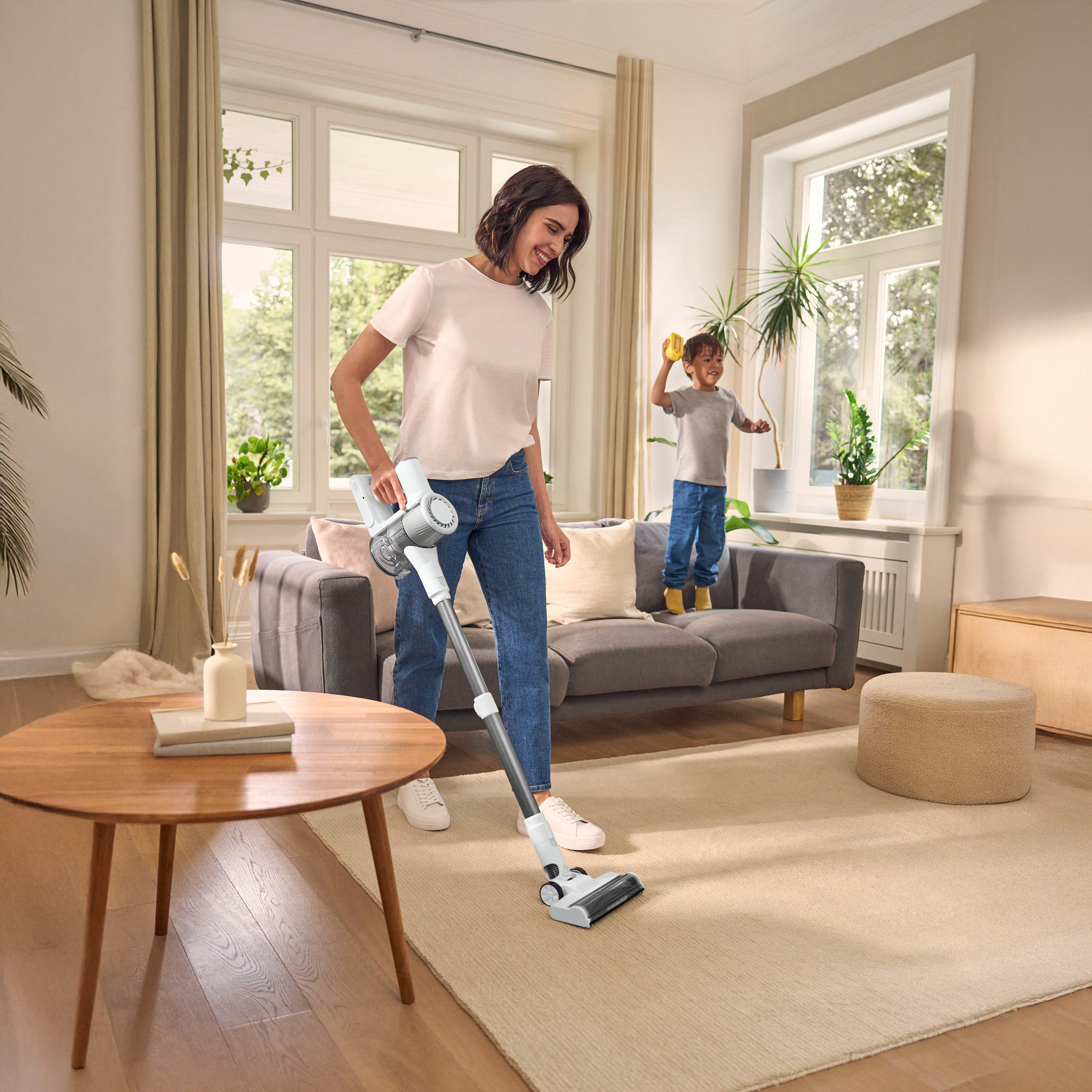 Frau staubsaugt Teppich mit Akkusauger, Kind steht auf Sofa. Runder Tisch links, Fenster im Hintergrund.