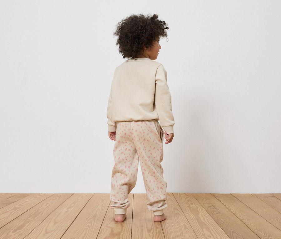 Ein Kind mit lockigem Haar steht mit dem Rücken zur Kamera und trägt einen beigen Pullover und eine Hose mit rosa Blumenmuster.