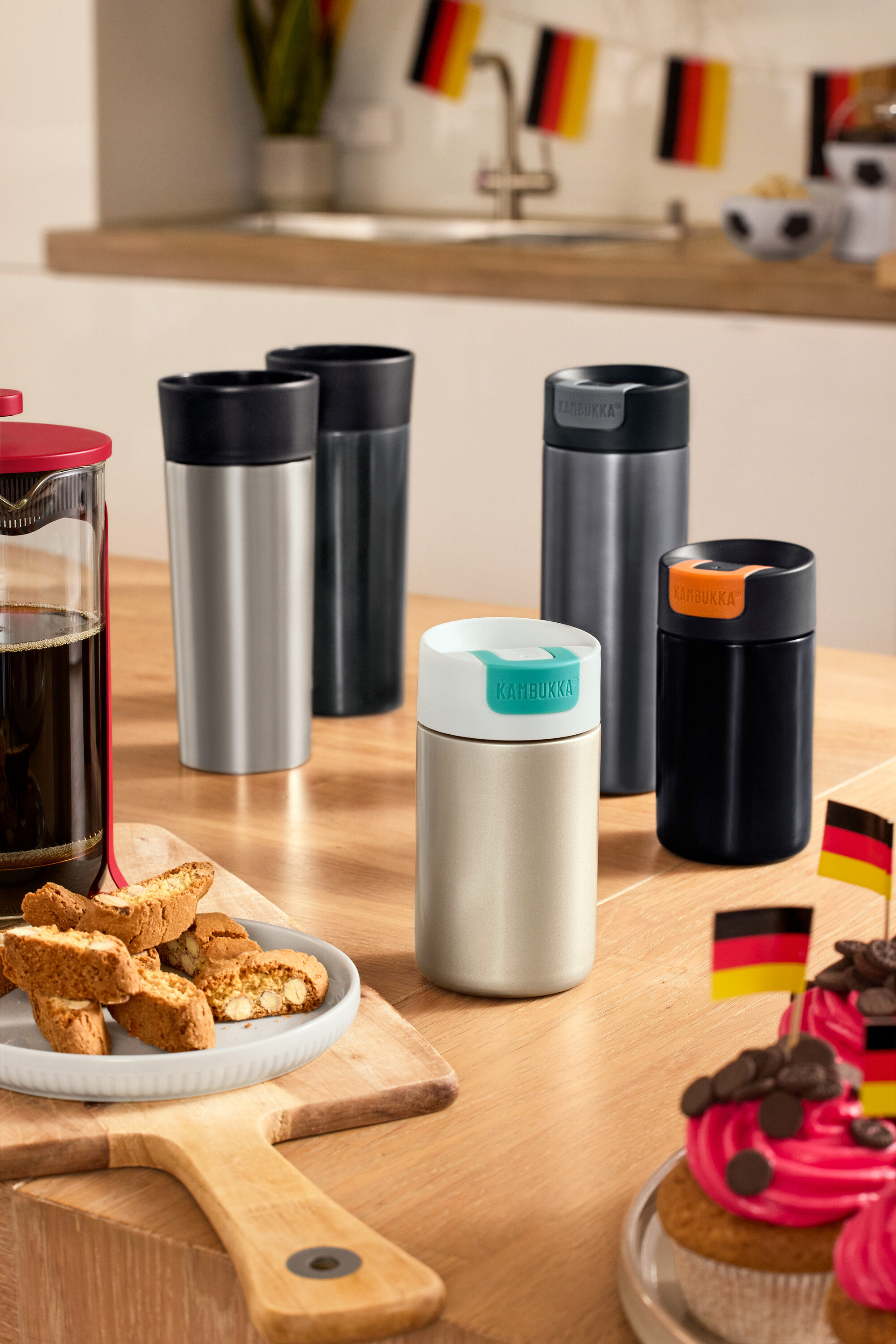 Auf einem Tisch stehen Coffee-to-go-Becher in Anthrazit und Edelstahl, Kambukka To-Go-Becher in Light Gold und Black Chrome sowie ein Kambukka To Go Becher in Grau.