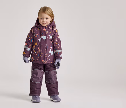 Ein kleines Mädchen steht in einem lila Schneeanzug mit Einhorn- und Regenbogenmuster und grauen Handschuhen und Schuhen vor einem hellen Hintergrund.
