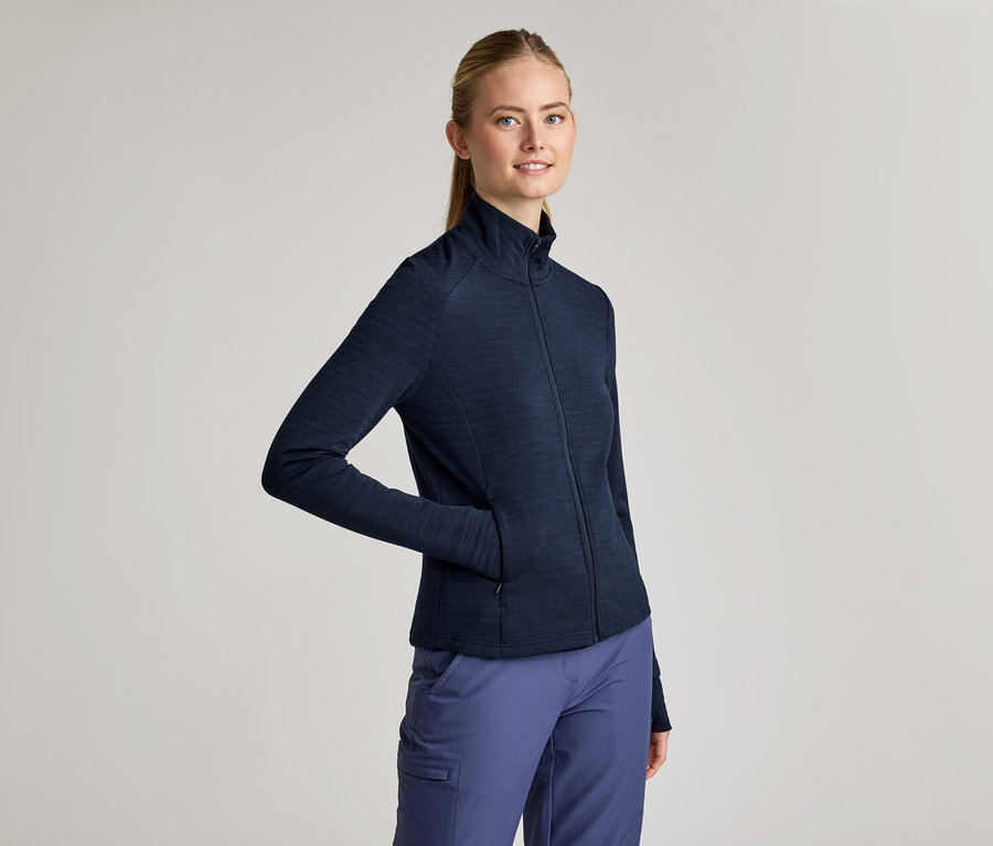Frau mit Zopf trägt eine marineblaue Jacke mit Reißverschluss und marineblaue Hose. Eine Hand steckt in der Jackentasche.