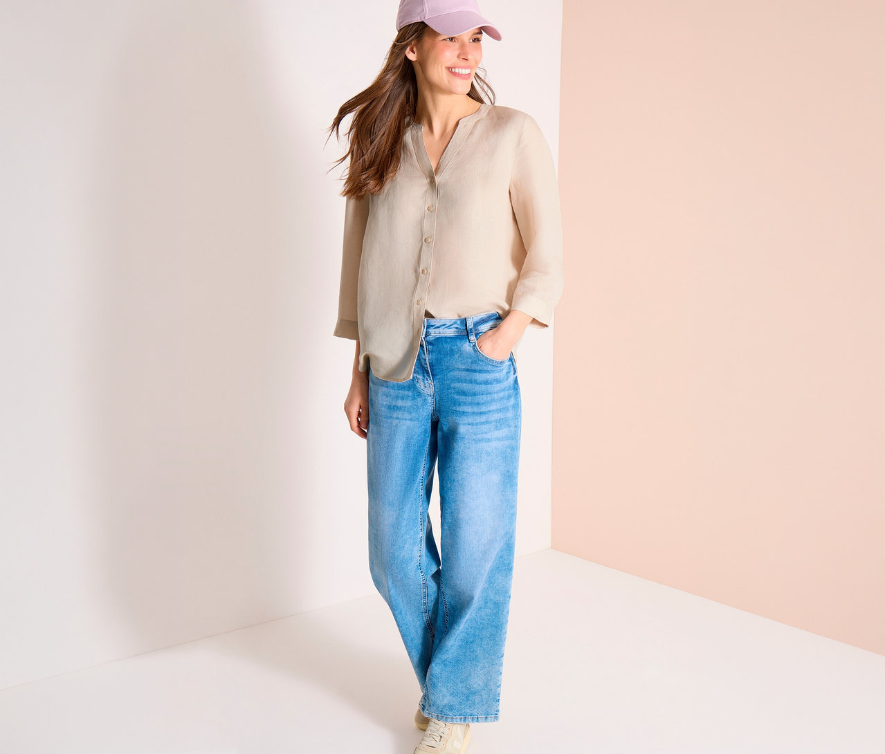 Lächelnde Frau mit rosa Kappe, beiger Bluse und blauen Jeans mit einer Hand in der Tasche.