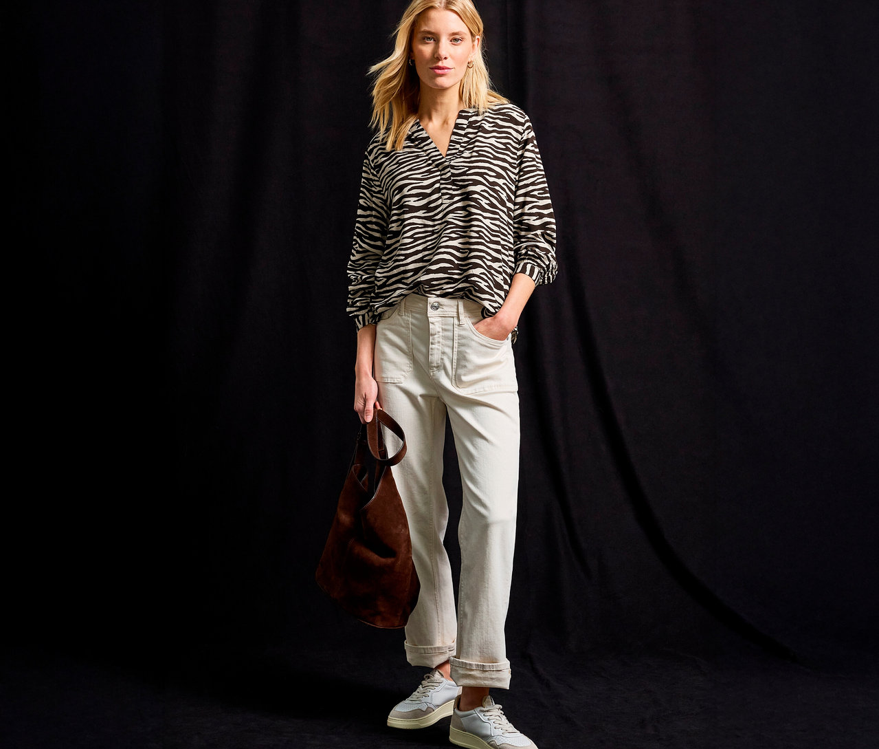 Blonde Frau trägt eine Street One Bluse mit Zebramuster, helle Hose, Sneaker und eine braune Tasche vor schwarzem Hintergrund.