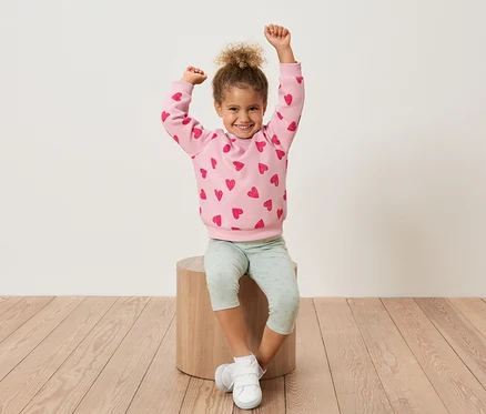 Ein junges Mädchen sitzt auf einem Holzhocker, die Arme hochgestreckt, und trägt ein rosa Sweatshirt mit roten Herzen und hellgrüne Leggings.