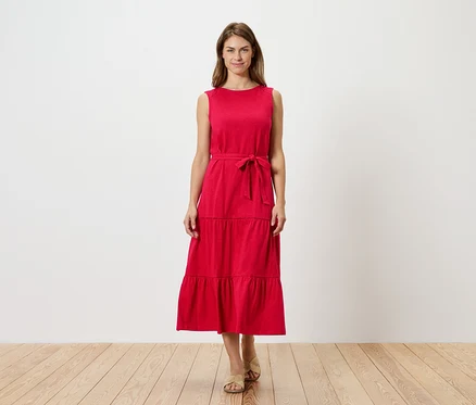 Eine Frau steht vor einer weißen Wand und trägt ein rotes, ärmelloses, gestuftes Kleid mit Gürtel und Sandalen.