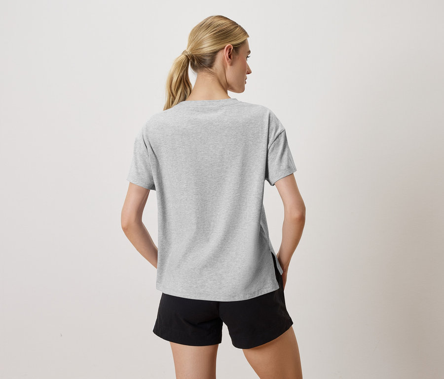Rückansicht einer Frau mit blonden Haaren, die ein graues T-Shirt und schwarze Shorts trägt und ihre Hände in den Hosentaschen hat.
