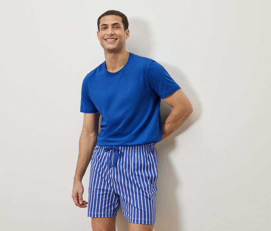 Mann mit blauem T-Shirt und gestreiften Shorts steht vor einer weißen Wand.