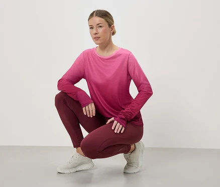 Frau hockt in Sportbekleidung, bestehend aus einem pink-roten, langärmligen Oberteil, weinroten Leggings und hellgrauen Turnschuhen vor einem weißen Hintergrund.