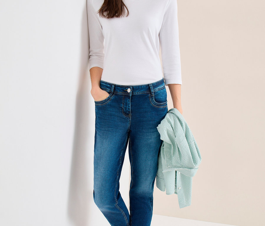 Frau in weißem Langarmshirt, blauer Baseballkappe und CECIL Jeans, lehnt an einer Wand und hält ein hellgrün gestreiftes Hemd.