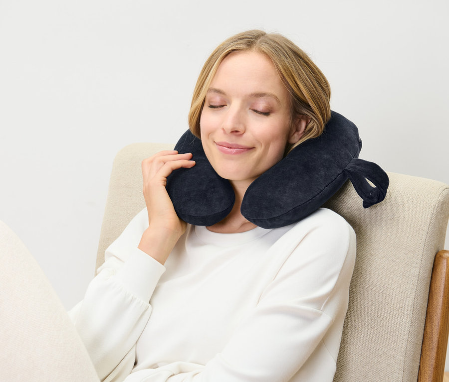 Frau sitzt mit geschlossenen Augen in einem Stuhl und trägt ein dunkelblaues Nackenkissen.