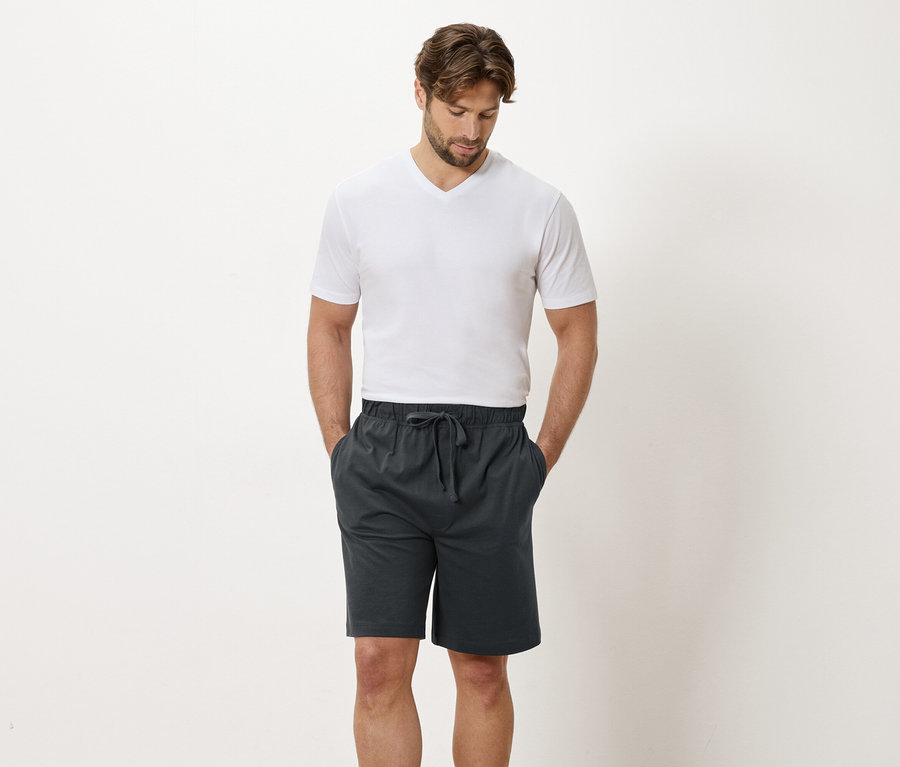 Mann mit weißem T-Shirt und grauen Shorts steht vor einer weißen Wand.
