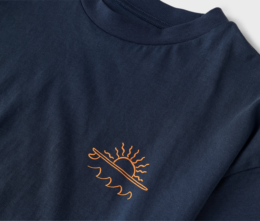 Detailaufnahme eines dunkelblauen T-Shirts mit orangefarbenem Aufdruck einer Sonne über einem Surfbrett und Wellen.