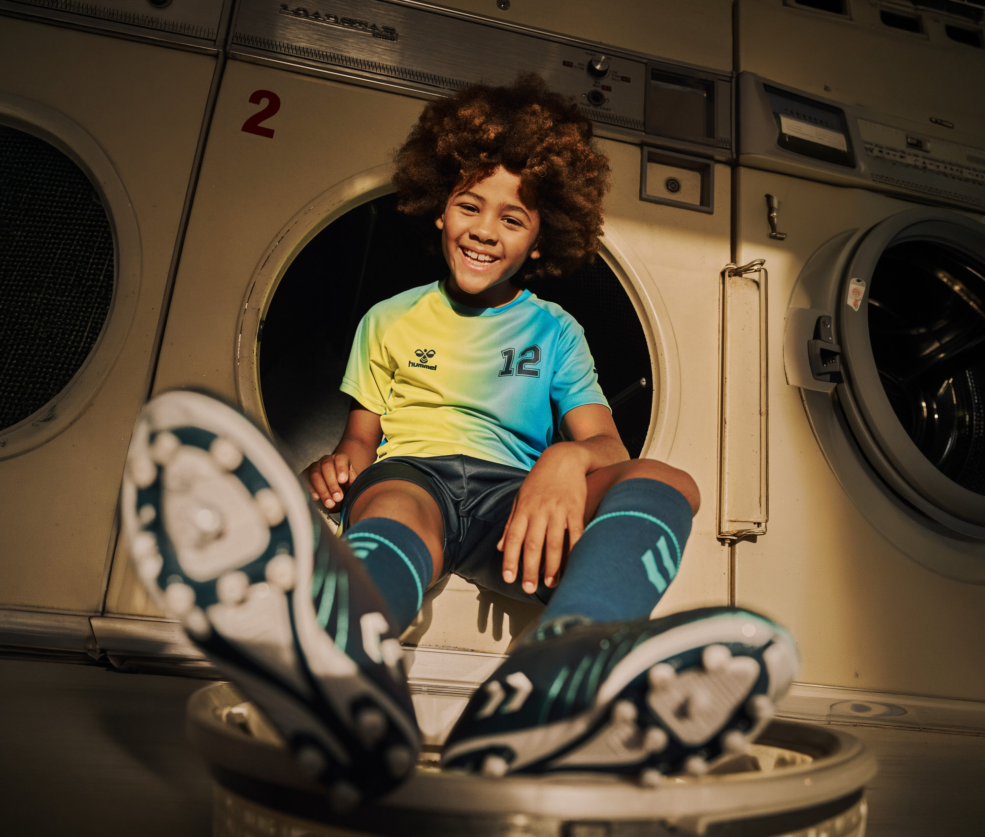 Ein lachender Junge mit Afro-Frisur sitzt in einer Waschmaschine und trägt ein gelb-blaues Trikot mit der Nummer 12, Shorts, Stutzen und Fußballschuhe.