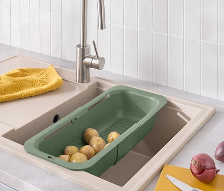 Kartoffeln in einem grünen Abtropfsieb, das in ein Spülbecken eingesetzt ist, neben einem gelben Tuch, einem gelben Schneidebrett und einer Zwiebel.