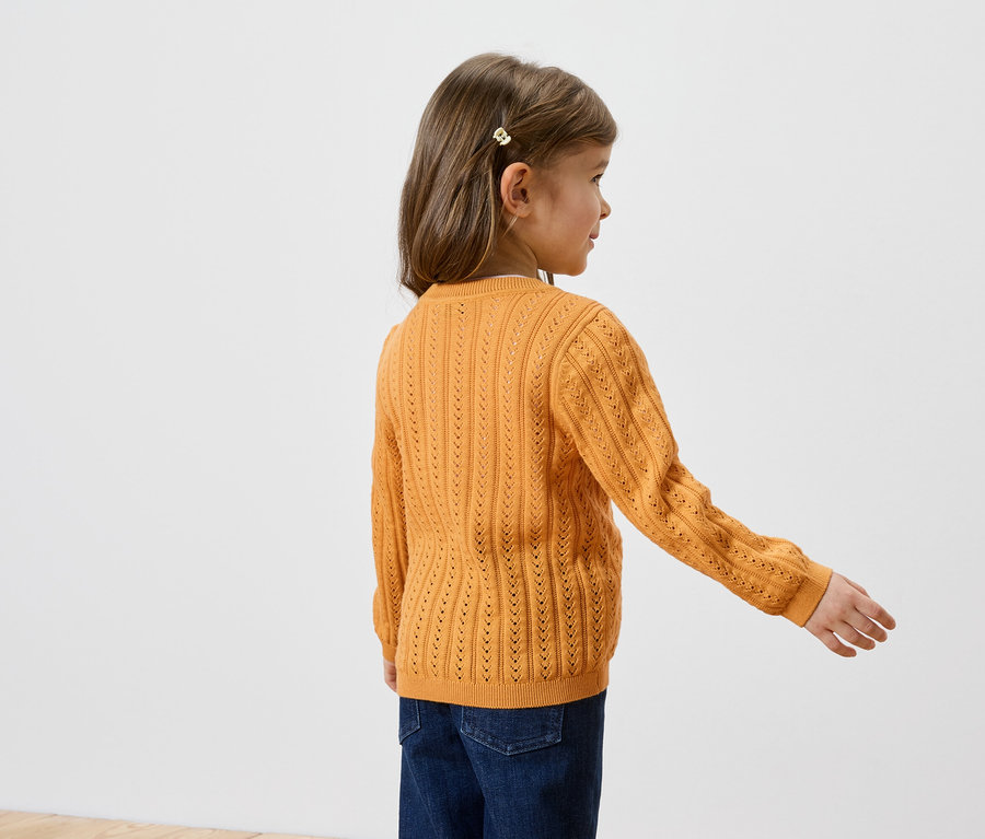 Ein junges Mädchen mit Haarspange trägt einen ockergelben Strickpullover und Jeans, von hinten gesehen.