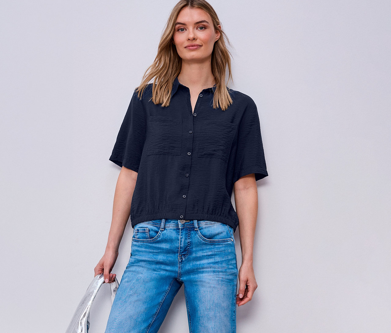 Frau trägt blaue Jeans und eine Street One Hemdbluse. Sie hält eine silberne Tasche.