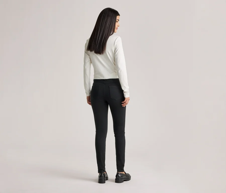 Eine Frau mit langen, dunklen Haaren, von hinten gesehen, trägt ein weißes Langarmshirt, schwarze Jeans und schwarze Schuhe.