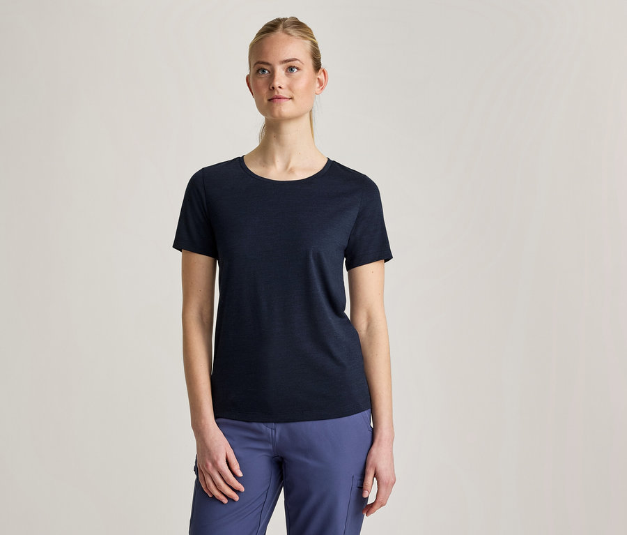 Eine Frau mit zurückgebundenem blondem Haar trägt ein dunkelblaues Kurzarm-T-Shirt und blaue Hosen, während sie vor einem hellgrauen Hintergrund steht.
