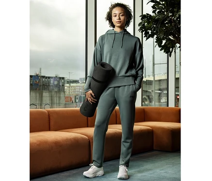 Frau in einem salbeifarbenen FILA Damen-Hoodie aus Modalmix und passender Jogginghose, die eine schwarze Yogamatte hält, steht in einem modernen Raum mit großen Fenstern.