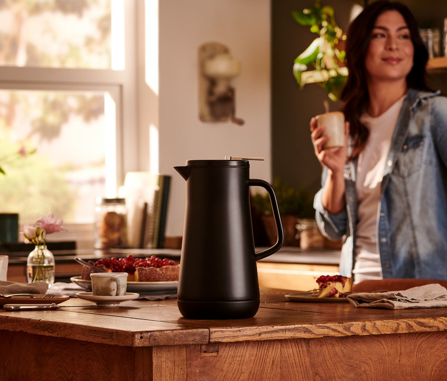 Schwarze Isolierkanne (1 l) steht auf einem Holztisch; Frau im Hintergrund hält eine Tasse.