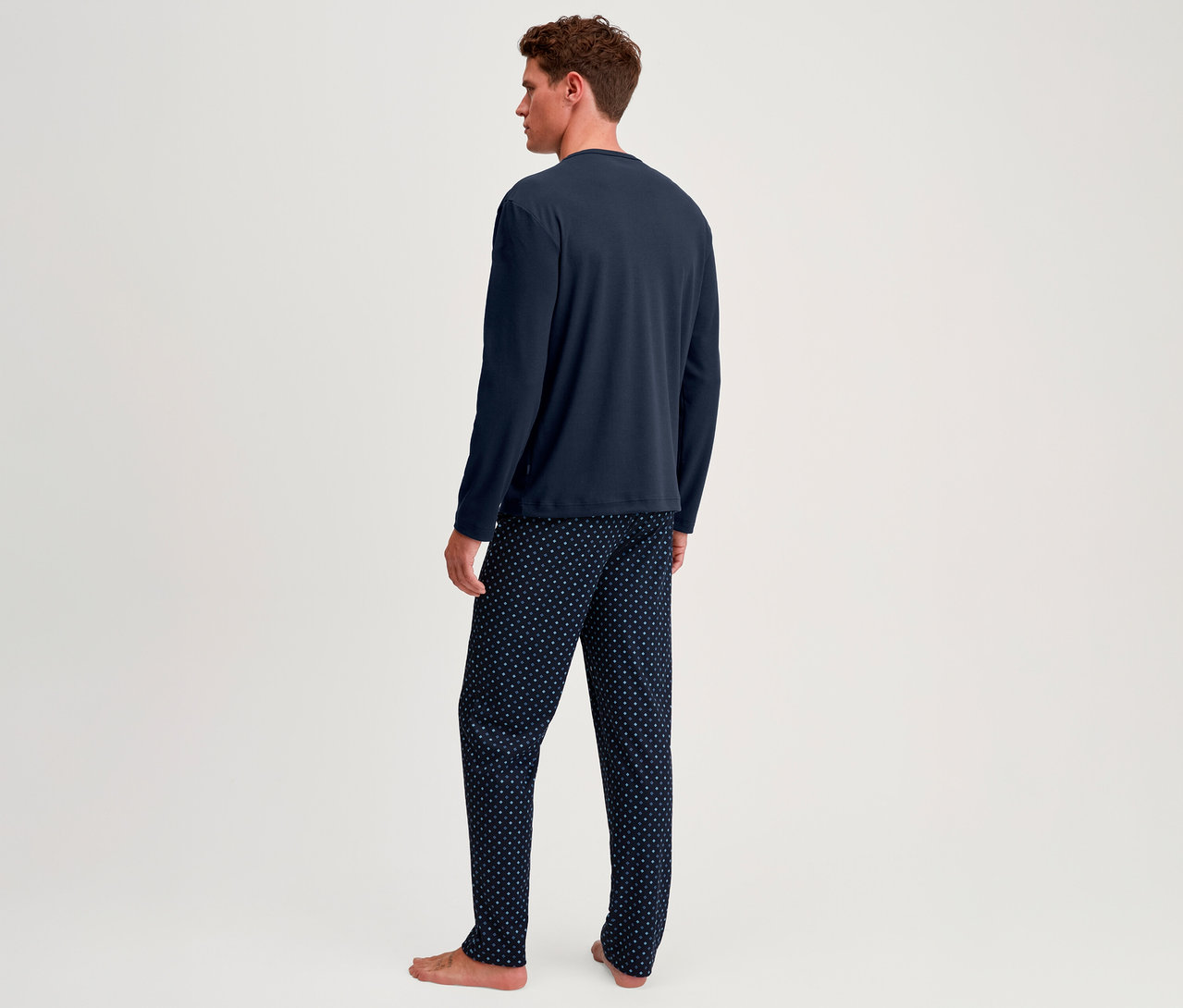 Mann von hinten, trägt ein dunkelblaues Langarmshirt und eine dunkelblaue Pyjamahose mit weißen Punkten, barfuß stehend.