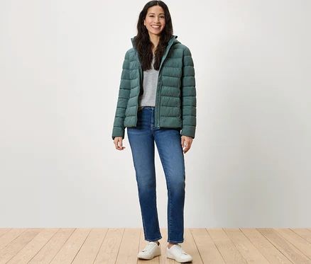 Eine Frau steht und lächelt in einer grünen Daunenjacke, blauen Jeans und weißen Turnschuhen.