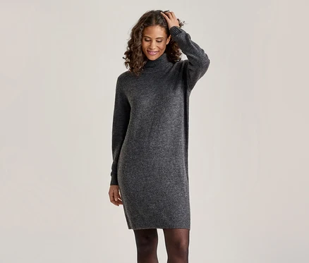 Eine lächelnde Frau mit lockigem Haar trägt ein dunkelgraues Rollkragen-Strickkleid und schwarze Strumpfhosen, eine Hand berührt ihr Haar.
