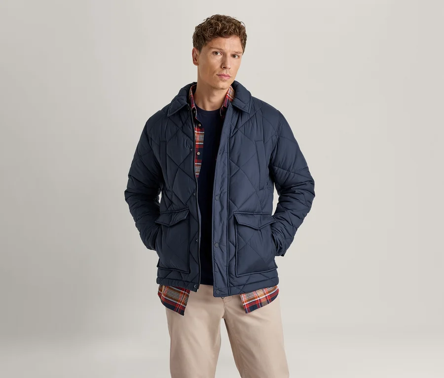 Ein Mann mit kurzen braunen Haaren steht frontal und trägt eine marineblaue Steppjacke über einem dunkelblauen Pullover und einem rot-blauen Karoshirt, dazu helle beigefarbene Hosen. Seine Hände stecken in den Jackentaschen.