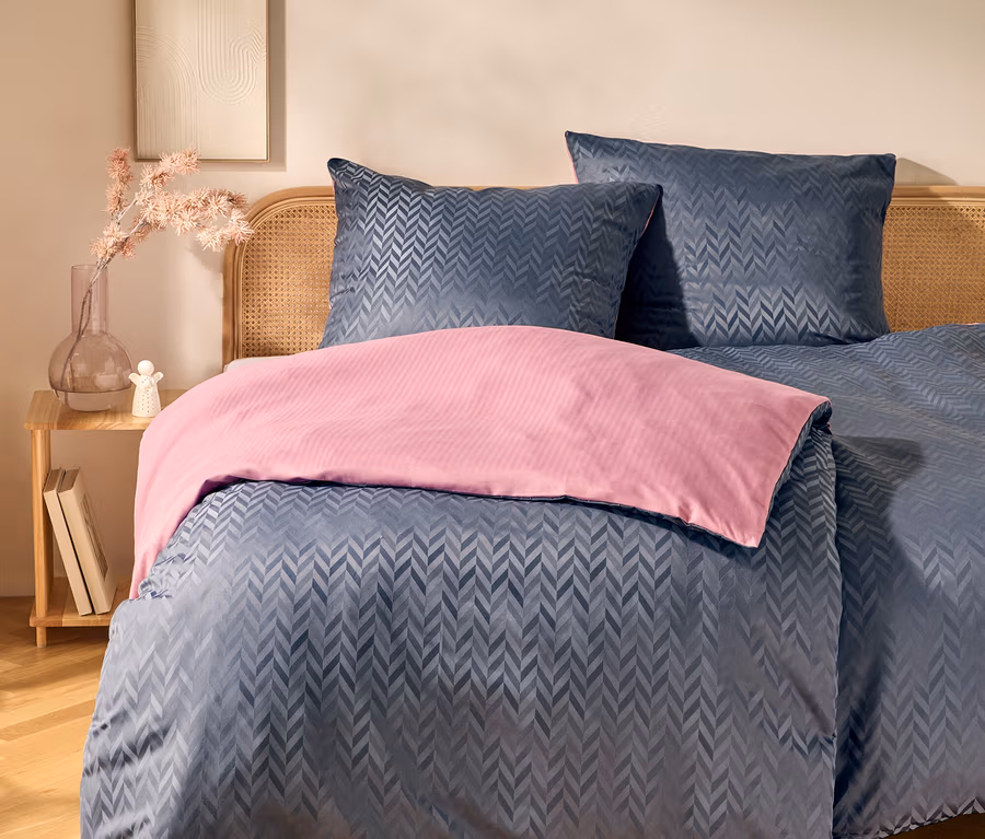Bett mit Rattan-Kopfteil, dunkelblauer Bettwäsche mit Chevron-Muster und rosa Überwurf. Daneben ein Holztisch mit Vase, rosa Blumen und Büchern. An der Wand hängt ein gerahmtes Kunstwerk.