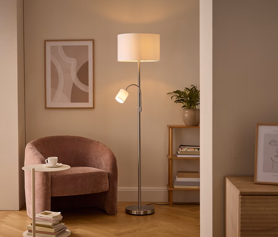 Gemütliche Wohnzimmerecke mit einer silbernen Stehlampe mit Haupt- und Leselicht, einem rosa Polstersessel und einem Beistelltisch mit Tasse. Wandkunst und ein Holzregal mit Pflanze sind ebenfalls sichtbar.