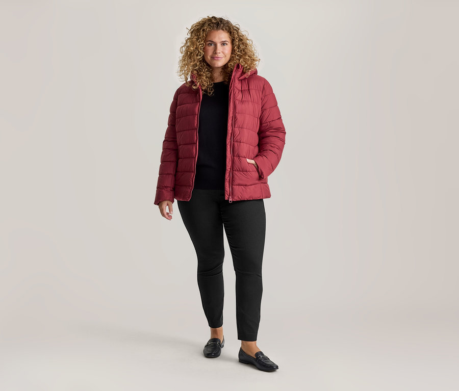 Frau mit lockigem Haar, roter Steppjacke, schwarzem Oberteil und schwarzer Stretchhose.