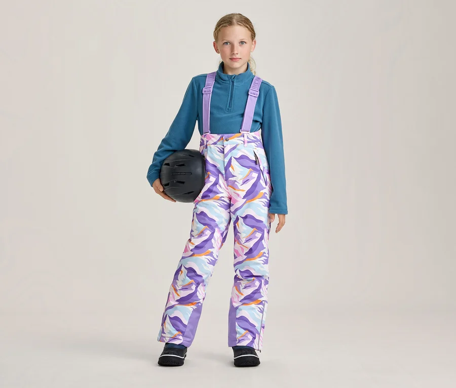 Junges Mädchen in einem petrolblauen Fleece-Oberteil und bunten gemusterten Skihosen mit lila Hosenträgern, das einen schwarzen Skihelm in der linken Hand hält.