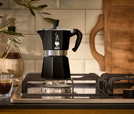 Ein schwarzer Espressokocher steht auf einem Gasherd neben einem Glas Espresso.
