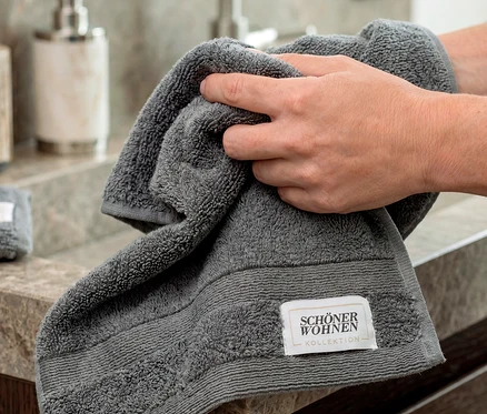 Hände halten ein anthrazitfarbenes SCHÖNER WOHNEN-Kollektion Gästehandtuch über einem Waschbecken.