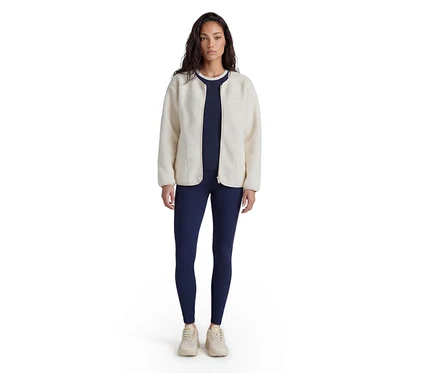 Frau steht, trägt eine cremefarbene Fleecejacke über einem marineblauen Oberteil und marineblauen Leggings, dazu helle Sneaker.