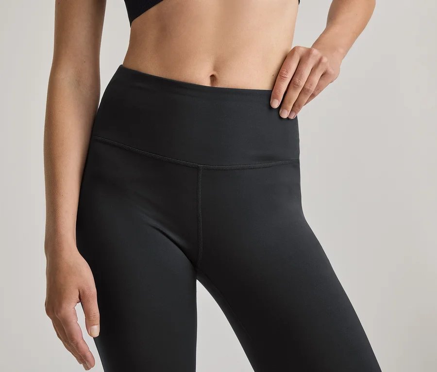 Nahaufnahme des Oberkörpers einer Person, die schwarze High-Waist-Leggings und einen schwarzen Sport-BH trägt. Die rechte Hand liegt auf der Hüfte.