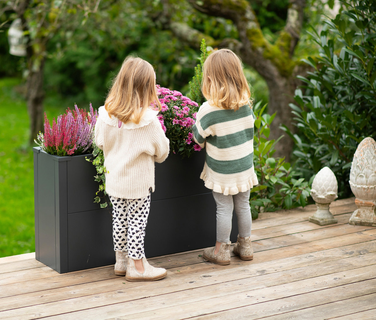 Zwei kleine Kinder stehen mit dem Rücken zum Betrachter auf einer Holzterrasse und blicken in einen anthrazitfarbenen Pflanzkasten mit Einlegeboden, der mit lila Heidekraut und rosa Blumen bepflanzt ist.