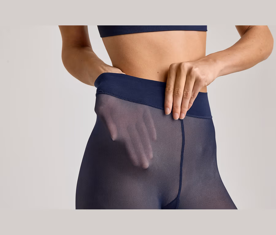 Nahaufnahme einer Frau in marineblauen, transparenten Strumpfhosen und passendem Sport-BH. Ihre Hände ziehen am Bund der Strumpfhose, wobei eine Hand durch den transparenten Stoff an ihrer Hüfte sichtbar ist.