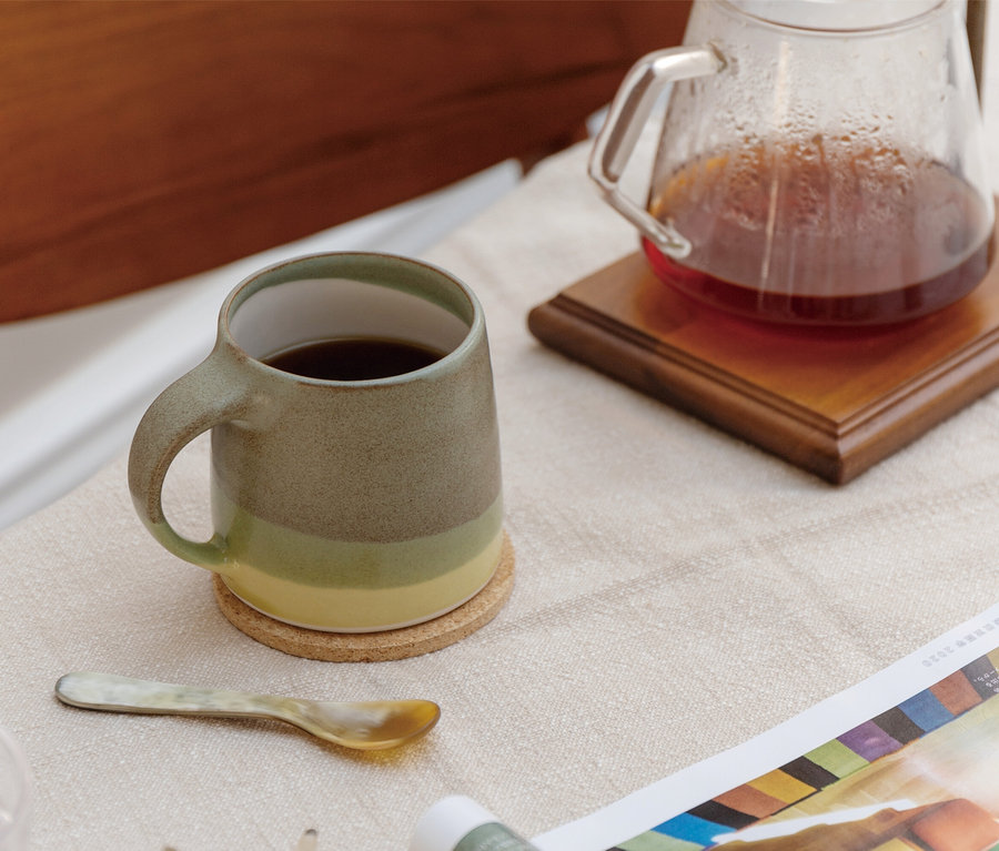 Keramiktasse mit grün-gelbem Farbverlauf und Kaffee auf Korkuntersetzer, daneben Glaskaraffe mit dunklem Getränk auf Holzuntersetzer. Ein kleiner Zierlöffel und ein farbiges Magazin liegen auf dem Tisch.
