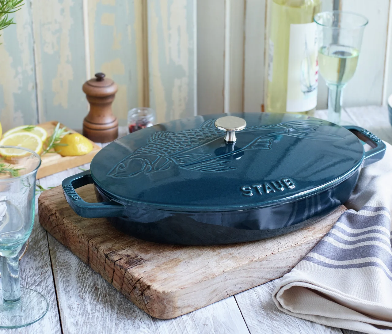 Dunkelblauer ovaler Gusseisentopf mit Fischmotiv auf dem Deckel, auf einem Holzbrett stehend. Zitronen, Rosmarin, eine Weinflasche und Gläser sind auf einem hellen Holztisch darum herum arrangiert.