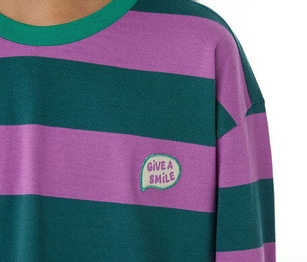 Nahaufnahme eines Kindes in einem SCHIESSER Kinder-Pyjama. Das Oberteil hat lila und dunkelgrüne Querstreifen, einen grünen Kragen und eine aufgestickte Sprechblase mit "GIVE A SMILE" auf der Brust.