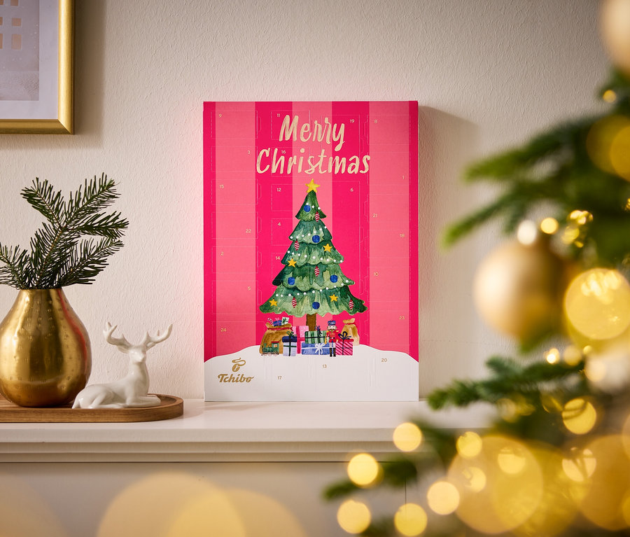 Rosa RA FL/OD Tchibo Adventskalender mit "Merry Christmas" und Weihnachtsbaum-Illustration, daneben eine goldene Vase mit Tannenzweigen und ein weißes Hirsch-Figürchen.
